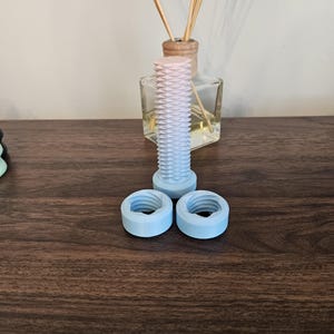 Op de afbeelding: Een 3D-geprint object in lichtblauw en roze, gevormd als een bout en twee moeren. Het object ligt op een houten oppervlak.
