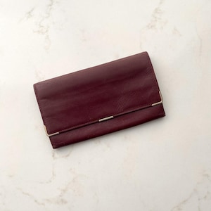 May include: A rectangular burgundy wallet with a smooth texture and a fold-over flap. The wallet features thin, metallic gold-toned corner accents on the bottom edges of the flap and a small, centred metallic gold-toned bar detail on the closure.