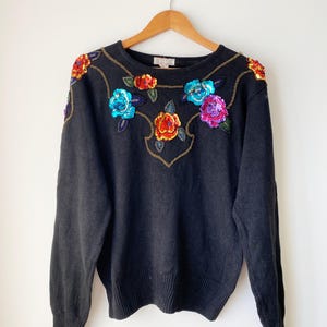 May include: Black sweater adorned with vibrant sequined floral embroidery. The flowers feature red, blue, orange, and purple hues, complemented by gold detailing. The sweater is displayed on a wooden hanger against a white backdrop.