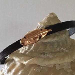 Puede incluir: Un brazalete de cuero negro con un dije de pluma dorada.