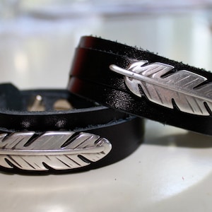 May include: Two black leather bracelets with silver feather charms. The bracelets are stacked on top of each other.