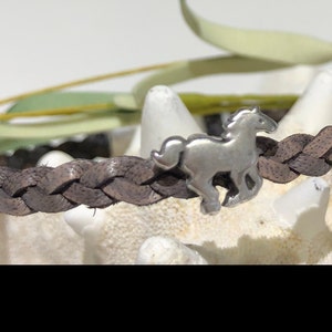 Puede incluir: Un brazalete trenzado de cuero marrón con un dije de caballo plateado.