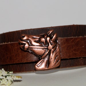May include: A brown leather wrap bracelet featuring a copper horse head design. The bracelet is adjustable and has a rustic look.