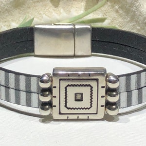 May include: A black and white striped leather bracelet with a silver square charm featuring a smaller square design inside. The bracelet has a magnetic clasp.