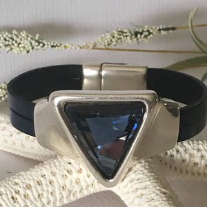 May include: A black leather bracelet with a silver triangle charm featuring a blue crystal. The bracelet has a magnetic clasp.