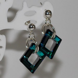 Può includere: Un paio di orecchini pendenti con un design a perno e anello argentato. Ogni orecchino presenta un cristallo di forma quadrata, color verde acqua. Gli orecchini sono esposti su un espositore bianco.