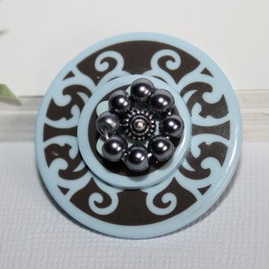 Peut inclure: Un bouton circulaire bleu et marron avec un motif décoratif et une fleur grise en perles au centre.