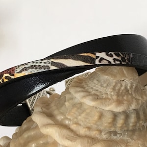 Op de afbeelding: Zwarte wikkelarmband met een band met dierenprints. De armband heeft een zilverkleurige sluiting en wordt gepresenteerd op een zeeschelp. Een modieus accessoire.