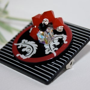 Peut inclure: Une broche carrée à rayures noires et blanches avec un motif circulaire rouge et blanc. Le cercle présente un motif floral blanc et un cristal avec des perles rouges.