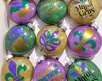 Puede incluir: Nueve adornos navideños de purpurina morada, verde y dorada con temas de Mardi Gras. Los adornos presentan texto como "Throw Me Something Mister" y "Laissez les bons temps rouler" e imágenes de flores de lis, máscaras y sombreros de bufón.