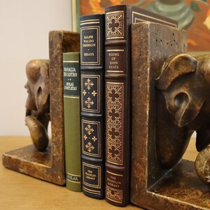 Puede incluir: Un par de sujetalibros con esculturas de elefantes con un acabado de madera marr&oacute;n. Los sujetalibros sostienen tres libros con letras doradas en los lomos. Los t&iacute;tulos de los libros son "Rosalia de Castro Obras Completas", "Ralph Waldo Emerson Essays" y "Poems of John Keats".
