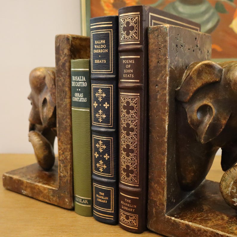 Puede incluir: Un par de sujetalibros con esculturas de elefantes con un acabado de madera marr&oacute;n. Los sujetalibros sostienen tres libros con letras doradas en los lomos. Los t&iacute;tulos de los libros son "Rosalia de Castro Obras Completas", "Ralph Waldo Emerson Essays" y "Poems of John Keats".