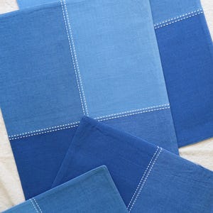 May include: Several rectangular blue placemats with a two-tone design. The top half is a lighter shade of blue, and the bottom half is a darker shade. White stitching accents the seam. The placemats are arranged on a white surface.