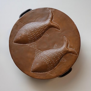 May include: A round, brown serving tray with two embossed fish designs. The fish have detailed scales and fins. The tray has two black handles on opposite sides. The background is a plain white.