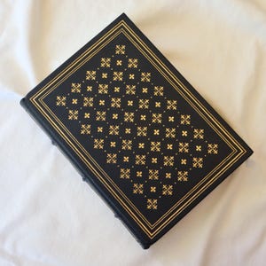 Puede incluir: Un libro de tapa dura negro con un dise&ntilde;o estampado en oro en la portada. El patr&oacute;n es un dise&ntilde;o geom&eacute;trico repetitivo de peque&ntilde;os cuadrados y cruces.