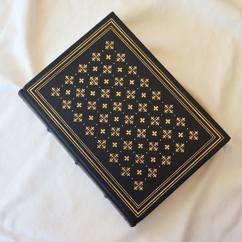 Puede incluir: Un libro de tapa dura negro con un dise&ntilde;o estampado en oro en la portada. El patr&oacute;n es un dise&ntilde;o geom&eacute;trico repetitivo de peque&ntilde;os cuadrados y cruces.
