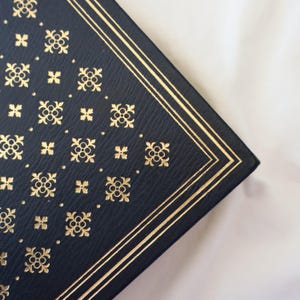 Puede incluir: Primer plano de la esquina de un libro de tapa dura de color azul oscuro con un borde dorado y un patr&oacute;n repetitivo de dise&ntilde;os florales dorados.