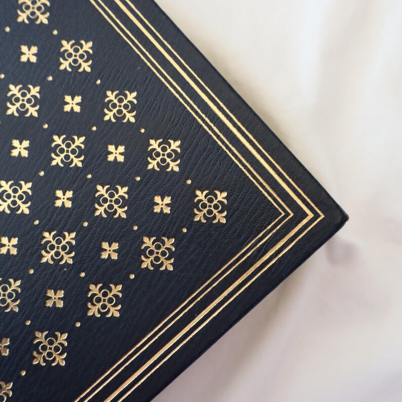 Puede incluir: Primer plano de la esquina de un libro de tapa dura de color azul oscuro con un borde dorado y un patr&oacute;n repetitivo de dise&ntilde;os florales dorados.