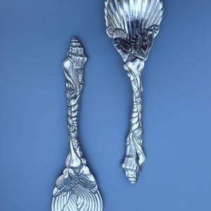 May include: Two silver-colored serving utensils with seashell designs. One is a serving spoon with a large shell-shaped bowl. The other is a serving fork with a split, fan-shaped bowl. Both have intricately detailed handles.