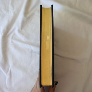 Puede incluir: El lomo de un libro de tapa dura con una cubierta negra y p&aacute;ginas doradas.