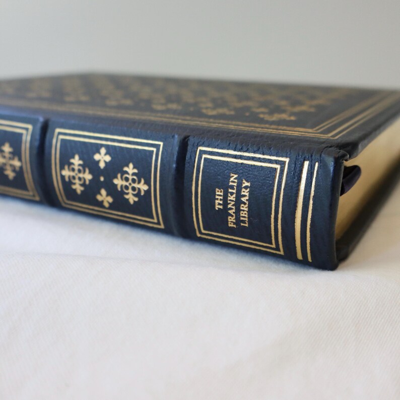 Puede incluir: Primer plano del lomo de un libro encuadernado en cuero negro con letras doradas que dicen "The Franklin Library".