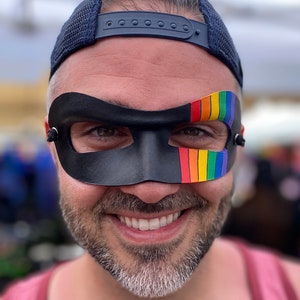 May include: A person wearing a black leather mask with a rainbow stripe on the right side. The mask covers the eyes and part of the nose. The person is smiling and wearing a blue baseball cap.