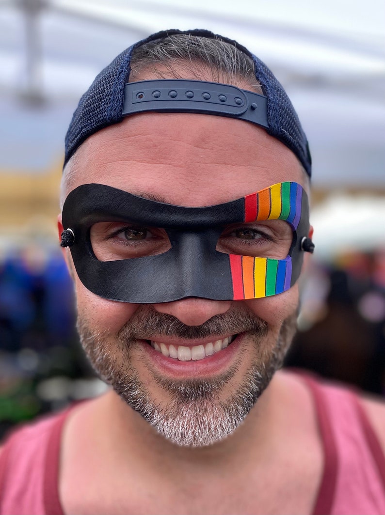 May include: A person wearing a black leather mask with a rainbow stripe on the right side. The mask covers the eyes and part of the nose. The person is smiling and wearing a blue baseball cap.