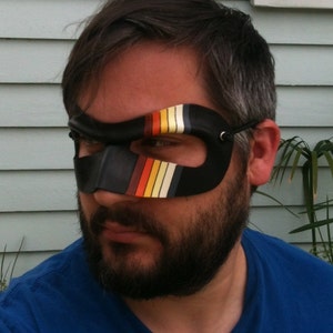 May include: A man wearing a black eye mask with colorful stripes. The mask has a black strap that goes around the back of the head. The man has a beard and is wearing a blue shirt.