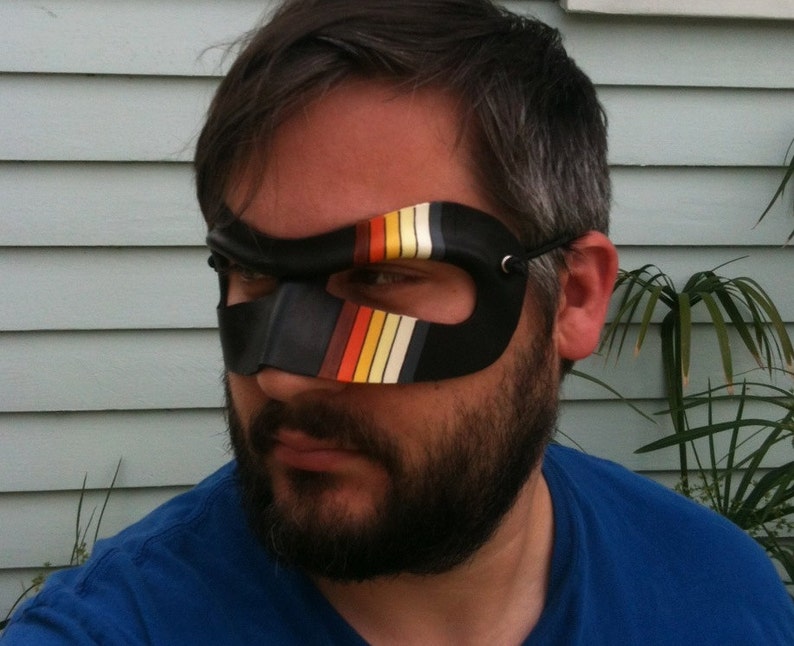 May include: A man wearing a black eye mask with colorful stripes. The mask has a black strap that goes around the back of the head. The man has a beard and is wearing a blue shirt.