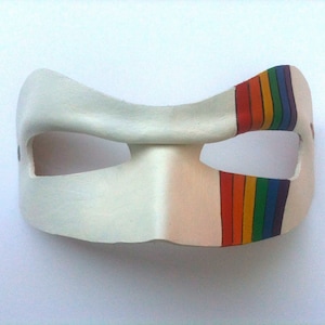 May include: A white leather mask with a rainbow stripe painted on the right side. The mask has two eye holes and is shaped like a crescent moon.