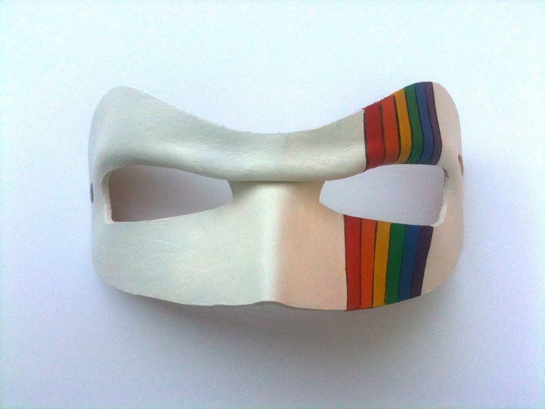 May include: A white leather mask with a rainbow stripe painted on the right side. The mask has two eye holes and is shaped like a crescent moon.