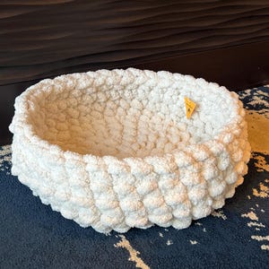 May include: A round, off-white, chunky knit basket with a thick, textured yarn construction. The basket features a small, triangular yellow tag attached to the inner rim. The design consists of large, interlocking loops of soft, plush material.