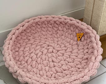 May include: A round, chunky knit pet bed in a soft pastel pink color. The bed features a thick, braided texture created from plush yarn. A small, triangular orange tag with black text is attached to the rim of the circular, bowl-shaped structure.