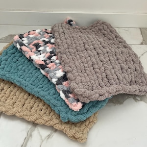 May include: A stack of four rectangular, chunky knit blankets made from thick, soft yarn. The blankets feature distinct colors including a solid beige, a solid teal, a variegated mix of pink, gray, and white, and a solid light brown top layer.