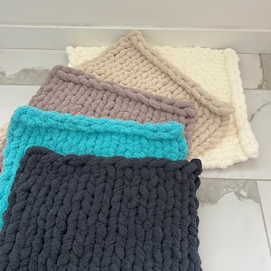 May include: A selection of chunky knit blankets in cream, tan, grey, teal, and dark grey. The blankets are square and have a thick, textured appearance. These soft, cosy blankets are ideal for home decor and measure approximately 76 cm x 76 cm.