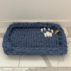 May include: A rectangular pet bed in a soft, chunky knit in a dusty blue colour. A white paw print design is on the bed's surface. A small orange tag is attached. The bed is designed for a small dog or cat.