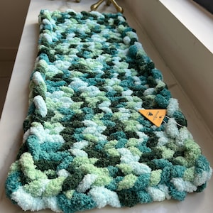 May include: A rectangular hand-knitted scarf featuring a chunky, textured pattern in a variegated color palette of dark forest green, medium teal, light mint green, and pale white yarn. A small triangular orange tag is attached to the side.