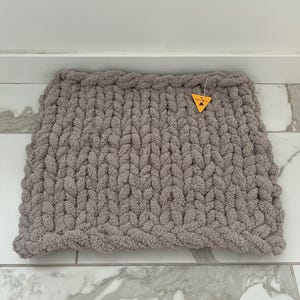 May include: A chunky knit, rectangular, grey blanket. The blanket is made of thick, soft yarn and has a textured appearance. A small, yellow tag is attached to the blanket. The blanket is displayed on a white and grey tiled surface.