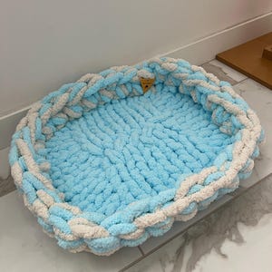 May include: A handmade pet bed in a light blue and cream color scheme. The bed is oval-shaped and made of thick, soft yarn. The edges are braided, and the interior is a solid light blue color. A small wooden tag is attached.
