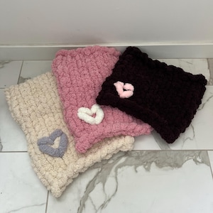 Puede incluir: Tres diademas de punto en crema, rosa y marrón oscuro, cada una adornada con un corazón aplicado. Las diademas están hechas de un hilo suave y texturizado, perfectas para añadir calidez y estilo.