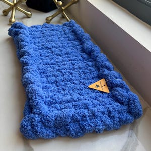 May include: A rectangular, bright blue crocheted blanket with a thick, textured, bobble-like border. A small, triangular, tan-colored paper tag is attached to the fabric, featuring the printed text "Mazzy & Ori" along with a small graphic icon.