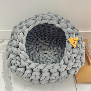 Pode incluir: Uma cama cinza para animais de estimação, tricotada à mão, com um design redondo em forma de caverna. A cama é feita de fio grosso e macio e apresenta uma textura tecida. Uma etiqueta amarela com o texto "Mazzy & Ori" pende de um lado.