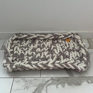 May include: A rectangular pet bed in a chunky knit design. The bed features a combination of grey and white yarn, creating a textured appearance. The edges are neatly finished with a braided border. A small tag is attached to the bed.
