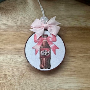 May include: A white round ornament with a pink bow and a Dr Pepper bottle graphic. The ornament is decorated with glitter.