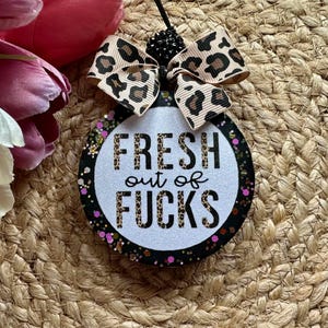 May include: A round hanging ornament with a white glittery center featuring the black text FRESH out of FUCKS in a leopard print pattern. The border is black with colorful glitter accents. A leopard print ribbon bow sits at the top with black beads.