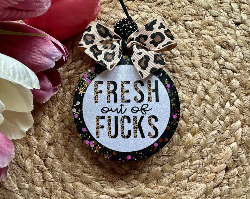 May include: A round hanging ornament with a white glittery center featuring the black text FRESH out of FUCKS in a leopard print pattern. The border is black with colorful glitter accents. A leopard print ribbon bow sits at the top with black beads.