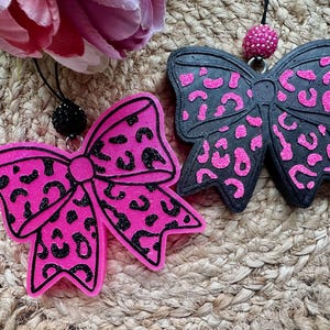 May include: Two bow-shaped ornaments featuring leopard print patterns. One bow is bright pink with black spots, and the other is black with bright pink spots. Each bow includes a small, round, textured bead attached to a black cord for hanging.
