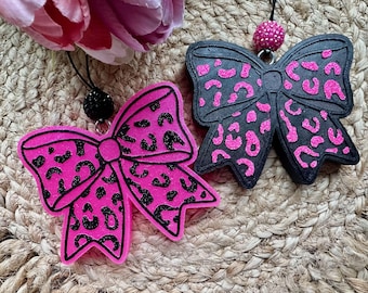 May include: Two bow-shaped ornaments with leopard print patterns. One bow is bright pink with black glitter spots, and the other is black with bright pink glitter spots. Each bow features a small, round, textured bead at the top attachment point.