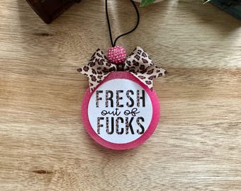 May include: A round, pink glittery air freshener hanging from a black elastic cord. It features a white center circle with the black text FRESH out of FUCKS in a leopard print pattern. A leopard print bow and a pink beaded accent sit on top.