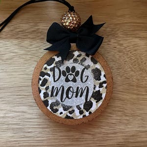 May include: A round, brown, wooden ornament with a black and white leopard print design. The ornament has a black ribbon and a gold glitter ball at the top. The ornament says "Dog Mom" with a paw print in the center.